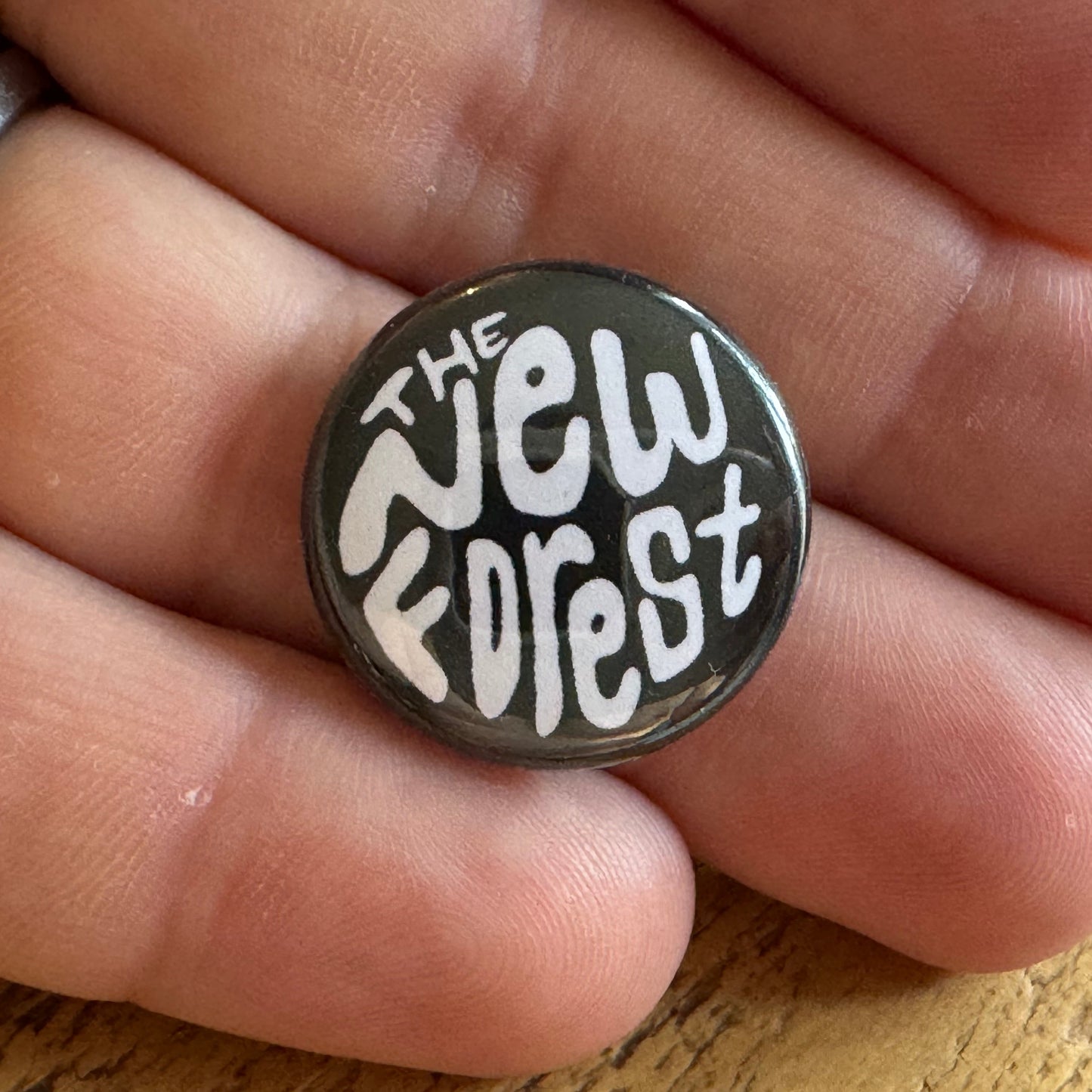 Button badge with 'The New Forest' text held between fingers on a wooden surface